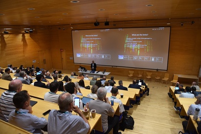 A group of people listening to a lecture with a speaker standing at the front and slides on a screen