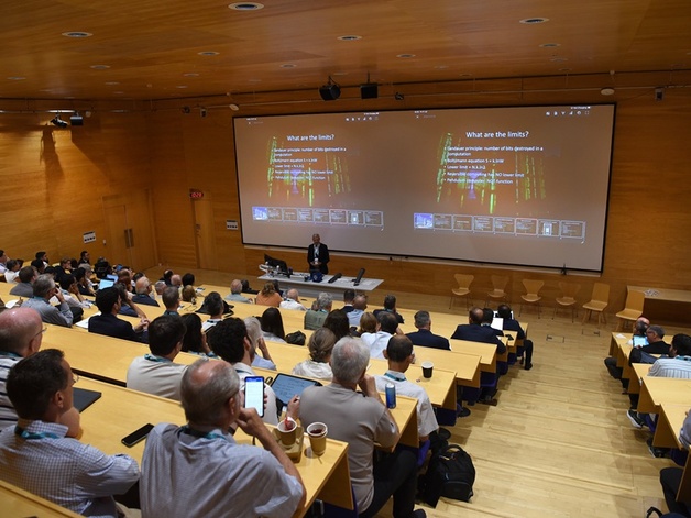 A group of people listening to a lecture with a speaker standing at the front and slides on a screen